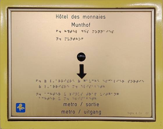 Brass sign at the Hôtel des Monnaies metro station in Brussels with printed French and Dutch text, lines of braille and a tactile arrow pointing to the exit