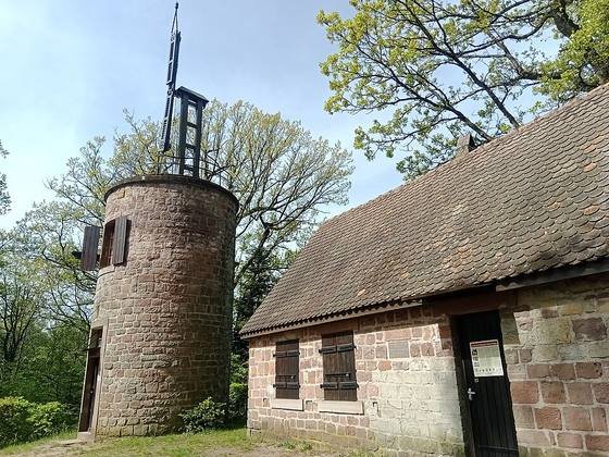 A round sandstone tower with a wooden mast and signal arms on top, next to a small stone house among trees
