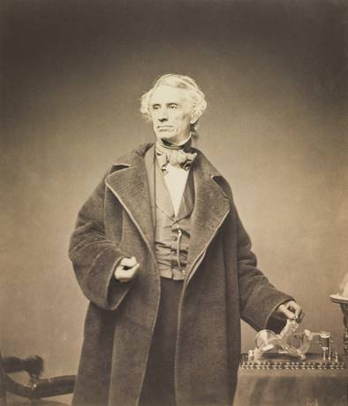 Sepia photograph of an elderly Samuel Morse with white hair, wearing a heavy overcoat and resting his hand on a telegraph register