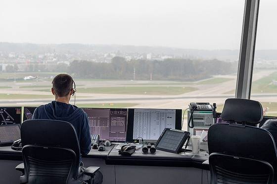An air traffic controller wearing a headset sits at screens in a glass-walled control tower overlooking a runway