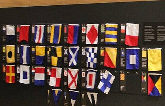 Museum wall display of rectangular international maritime signal flags in red, yellow, blue, black and white, each with a small label