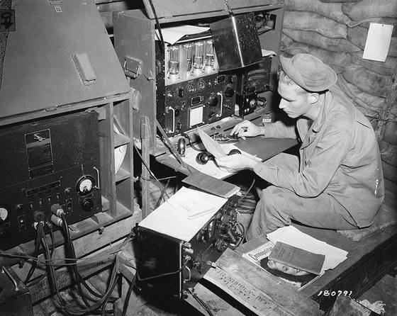 Black-and-white photo of a U.S. Army radio operator in uniform working at radio equipment with valves and a telegraph key in a sandbagged position