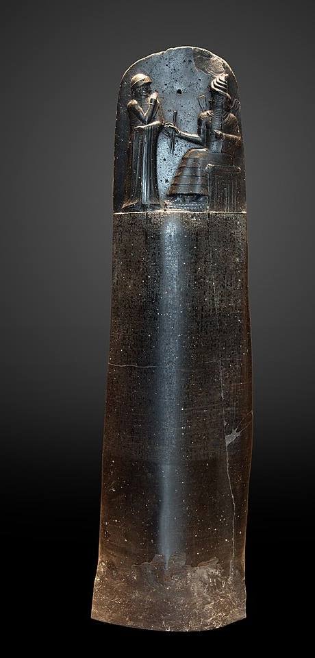 A tall black stone stele with a carved scene of a king before a seated god at the top and dense columns of cuneiform below